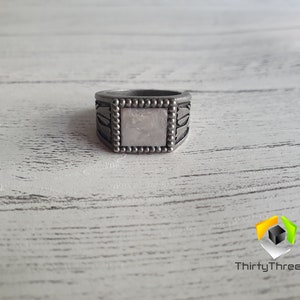 May include: A silver ring with a square, white stone set in the center. The ring has a beaded border and intricate designs on the sides.