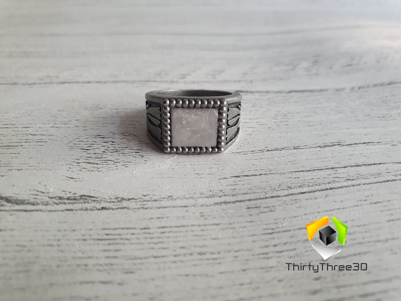 May include: A silver ring with a square, white stone set in the center. The ring has a beaded border and intricate designs on the sides.