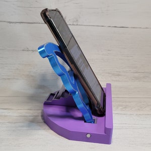 Peut inclure: Un support de t&eacute;l&eacute;phone violet avec un support de t&eacute;l&eacute;phone bleu. Le support a une base arrondie et une plate-forme sur&eacute;lev&eacute;e pour le t&eacute;l&eacute;phone. Le support de t&eacute;l&eacute;phone est en forme de lettre 'F'.