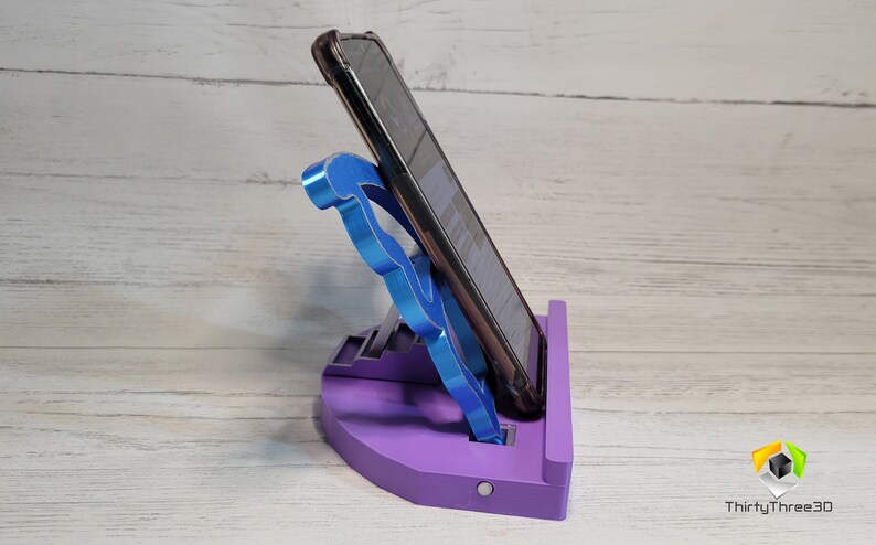 Peut inclure: Un support de t&eacute;l&eacute;phone violet avec un support de t&eacute;l&eacute;phone bleu. Le support a une base arrondie et une plate-forme sur&eacute;lev&eacute;e pour le t&eacute;l&eacute;phone. Le support de t&eacute;l&eacute;phone est en forme de lettre 'F'.