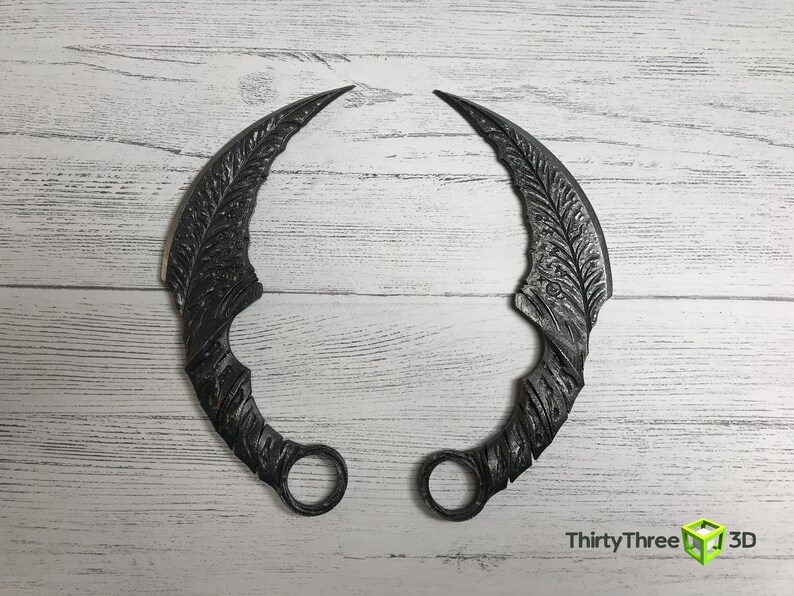 May include: A pair of black metal throwing knives with intricate designs. The knives are curved and have a ring at the end of the handle.