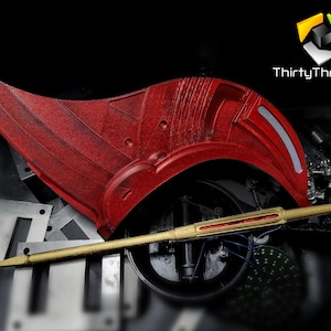 May include: A red and gold 3D printed prop, possibly a weapon or part of a costume. The prop has a curved, wing-like shape and a long, thin handle. The prop is resting on a black surface.