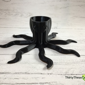 May include: A black octopus-shaped holder with eight tentacles. The holder has a round opening at the top, suitable for holding small items.