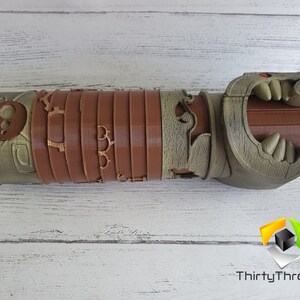 May include: A 3D printed, brown and green, detailed, articulated, dragon-like hand with a red eye. The hand is open and has sharp teeth.