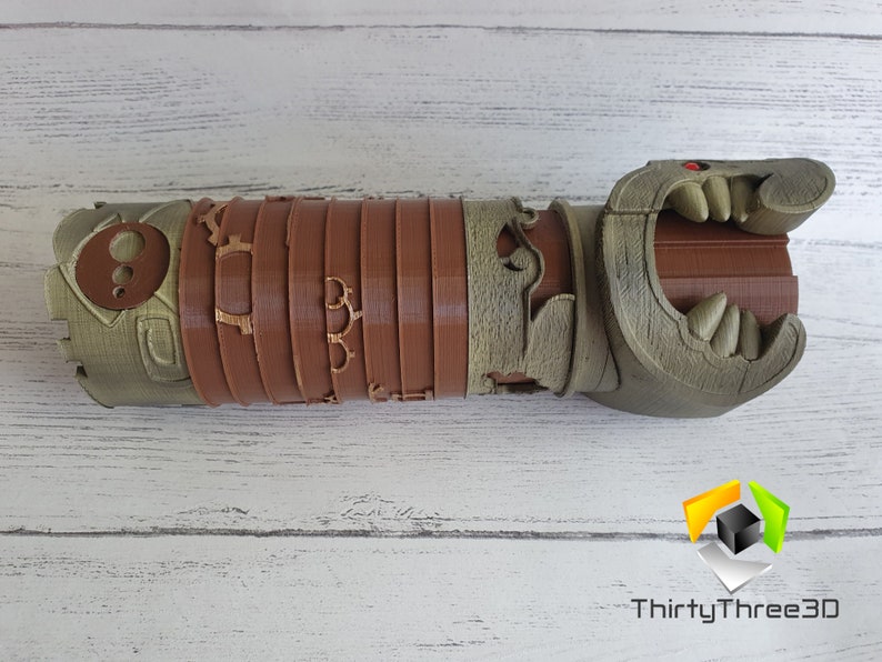 May include: A 3D printed, brown and green, detailed, articulated, dragon-like hand with a red eye. The hand is open and has sharp teeth.