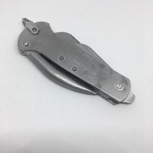 Puede incluir: Un cuchillo plegable plateado con una hoja dentada y un peque&ntilde;o agujero en el mango. El cuchillo est&aacute; cerrado y tiene una superficie texturizada.