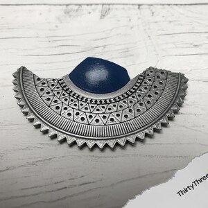May include: A silver-colored, geometric, half-circle pendant with a blue center. The pendant has a detailed design with a repeating pattern of triangles and circles.