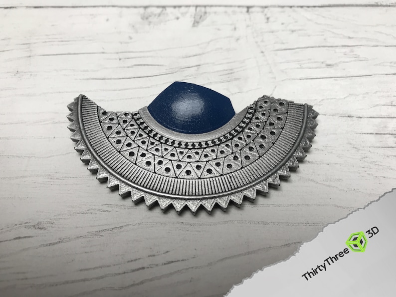 May include: A silver-colored, geometric, half-circle pendant with a blue center. The pendant has a detailed design with a repeating pattern of triangles and circles.
