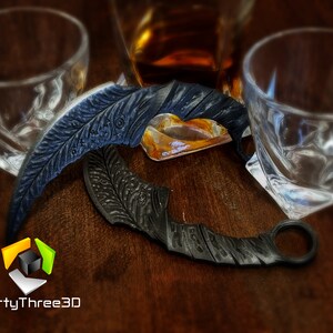May include: Two black metal throwing knives with intricate designs, one with a ring on the end, on a wooden surface with two empty glasses and a bottle of amber liquid.