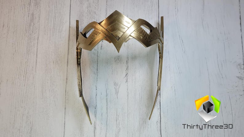 May include: A gold-colored metal crown with a geometric design. The crown is made of two pieces that are connected at the top. The crown is on a white wooden background.
