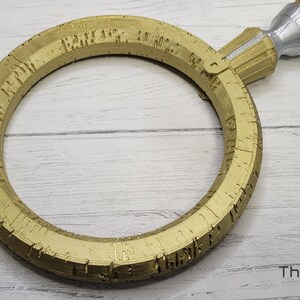 Pode incluir: Um anel dourado com detalhes intrincados e uma al&ccedil;a prateada. O anel &eacute; circular e tem uma superf&iacute;cie texturizada.