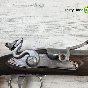 Puede incluir: Vista detallada de una r&eacute;plica de pistola de chispa. La pistola presenta una culata de madera marr&oacute;n oscuro, componentes met&aacute;licos de color plateado y detalles en color lat&oacute;n. El gatillo, el martillo y otros mecanismos son claramente visibles, mostrando el intrincado dise&ntilde;o.