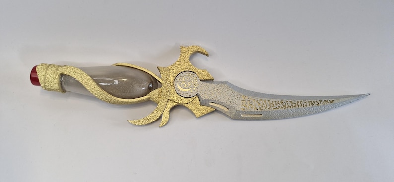 May include: An ornate dagger with a clear glass handle filled with a sand-like substance. The hilt and decorative elements are gold-colored, and the blade is silver with etched details. A red gem is at the end of the handle.