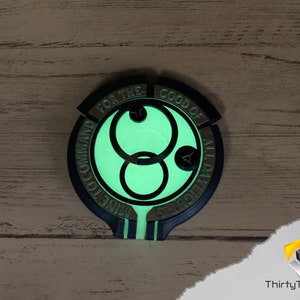May include: A black and gold circular pendant with a glowing green symbol in the center. The symbol is a double circle with a smaller circle in the middle. The text around the symbol reads "For the Good of All Mankind."