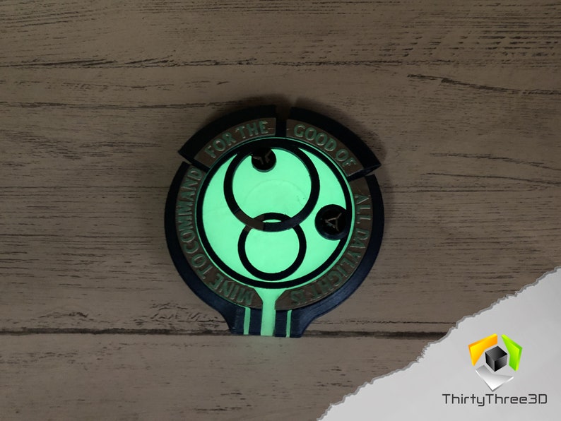 May include: A black and gold circular pendant with a glowing green symbol in the center. The symbol is a double circle with a smaller circle in the middle. The text around the symbol reads "For the Good of All Mankind."