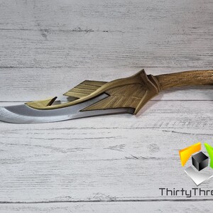 May include: A gold and silver fantasy-style dagger with a detailed handle and a sharp blade. The dagger is lying on a white wooden surface.