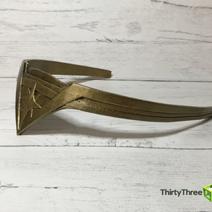 May include: A gold-colored headband with a star-shaped design. The headband is made of a smooth, hard plastic.