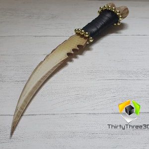 May include: A white and brown dagger with a serrated blade and a black leather handle with gold beads. The dagger is on a white wooden surface.