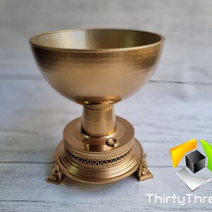 May include: A gold-colored metal bowl with a decorative base and three legs. The bowl is made of a smooth, shiny metal and has a round shape.