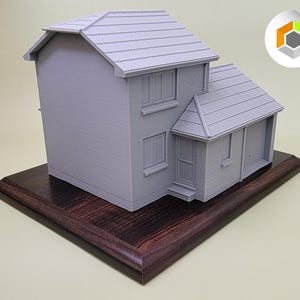 May include: A gray 3D printed model of a house on a dark wood base. The house has a garage and a small front porch.