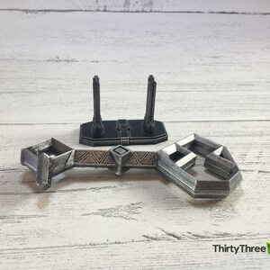 May include: A silver-colored 3D printed model of a bridge with two towers. The bridge is made of interlocking pieces and has a geometric design. The towers are tall and slender, with a decorative top.