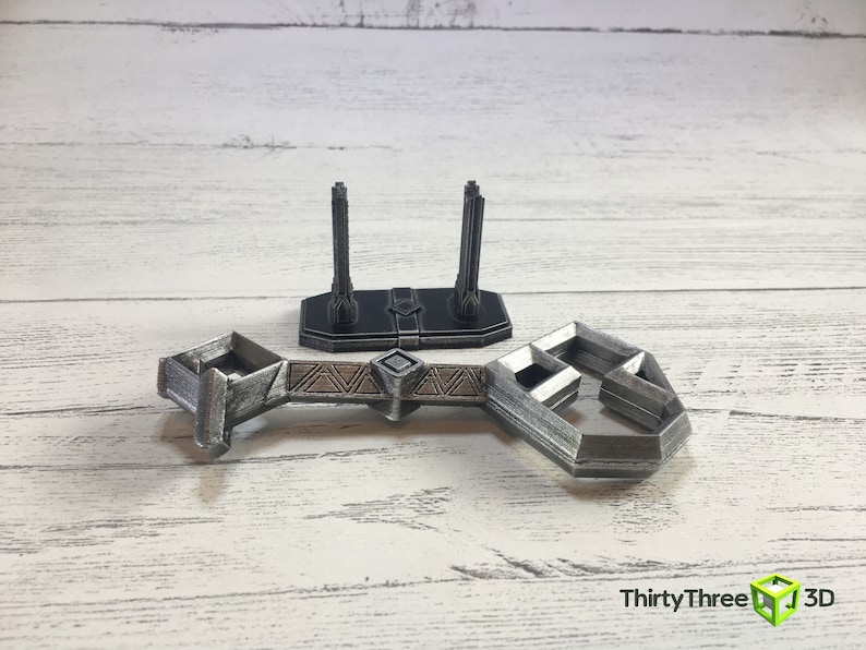 May include: A silver-colored 3D printed model of a bridge with two towers. The bridge is made of interlocking pieces and has a geometric design. The towers are tall and slender, with a decorative top.