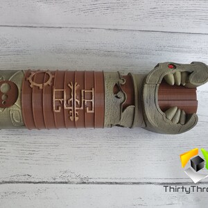 May include: A 3D printed, brown and green, detailed, articulated, dragon-like hand with a red eye. The hand is made of multiple sections that can be moved. The hand is on a white wooden surface.