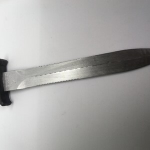Puede incluir: Un cuchillo de buceo con mango negro y hoja dentada. La hoja es de acero inoxidable y tiene una punta afilada.