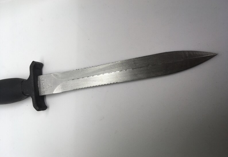 Puede incluir: Un cuchillo de buceo con mango negro y hoja dentada. La hoja es de acero inoxidable y tiene una punta afilada.