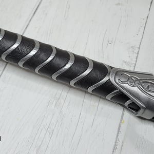 May include: Close-up of a black and silver handle with a leather grip and a silver metal cap with a decorative design. The handle is for a prop weapon.