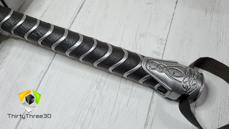 May include: Close-up of a black and silver handle with a leather grip and a silver metal cap with a decorative design. The handle is for a prop weapon.
