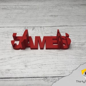 May include: Red 3D printed name sign spelling "JAMES" on a white wood background.