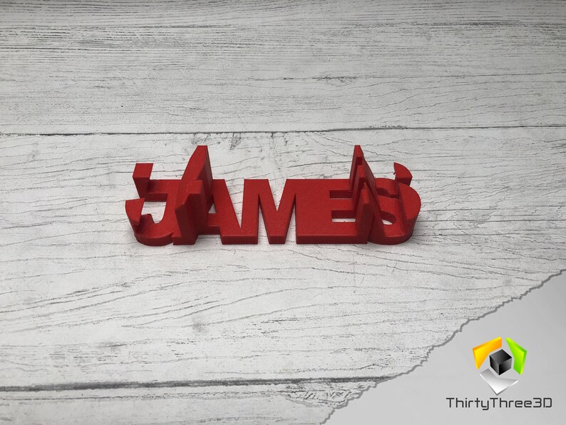 May include: Red 3D printed name sign spelling "JAMES" on a white wood background.