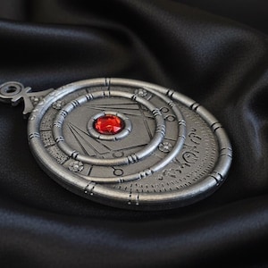 May include: A silver-colored pendant with a central red gemstone. The pendant features concentric circular designs and geometric patterns. The background is a dark, textured fabric, enhancing the pendant's details. The pendant has a loop at the top for a chain.