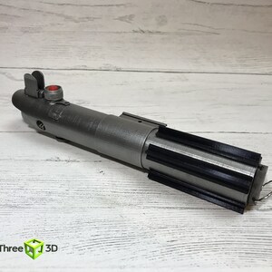 May include: A silver lightsaber prop with a black grip and a red button. The lightsaber is on a white wooden surface.