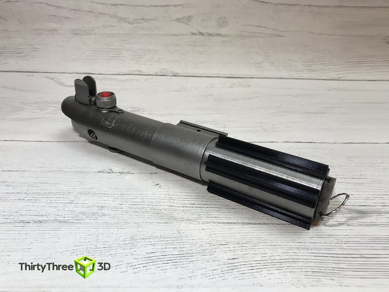 May include: A silver lightsaber prop with a black grip and a red button. The lightsaber is on a white wooden surface.