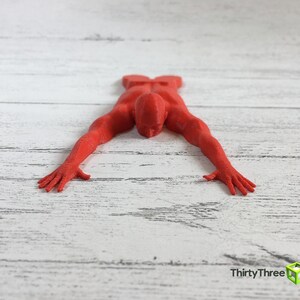 May include: A red 3D printed figurine of a person lying face down on a white wooden surface.