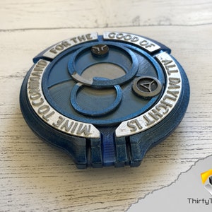 May include: A blue and silver 3D printed disc with the text "MINE TO COMMAND FOR THE GOOD OF ALL DYRLICHT IS" around the edge. The disc has a central design of three interlocking circles.