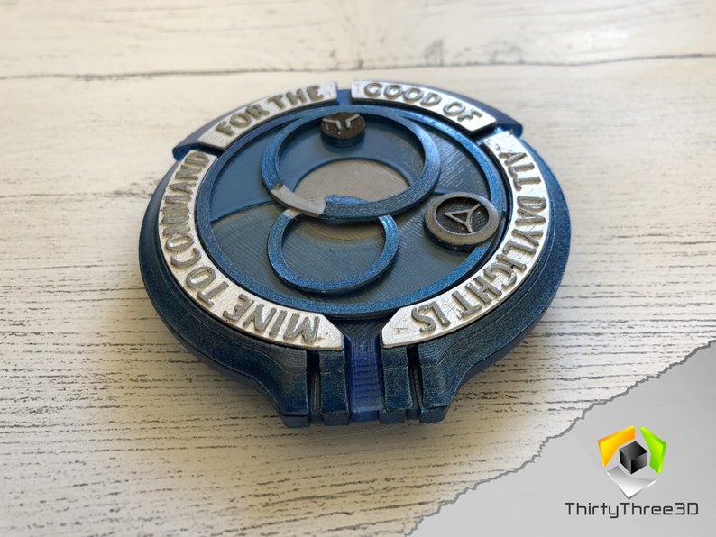 May include: A blue and silver 3D printed disc with the text "MINE TO COMMAND FOR THE GOOD OF ALL DYRLICHT IS" around the edge. The disc has a central design of three interlocking circles.