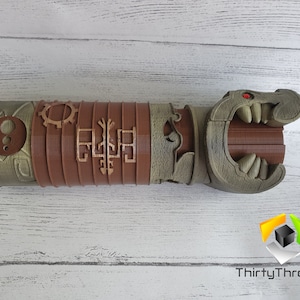 May include: A 3D printed, brown and green, detailed, articulated, dragon-like hand with a red eye. The hand is made of multiple sections that can be moved. The hand is on a white wooden surface.