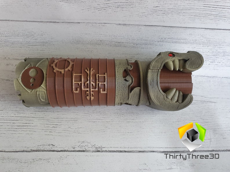 May include: A 3D printed, brown and green, detailed, articulated, dragon-like hand with a red eye. The hand is made of multiple sections that can be moved. The hand is on a white wooden surface.