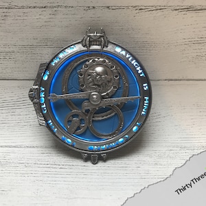 May include: A silver-colored metal circular pendant with a blue background and intricate gears. The pendant has the text "The Glory of Heaven" and "Daylight is Mine to Command" inscribed around the edge.