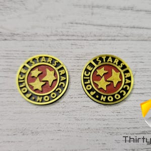 Puede incluir: Dos insignias de metal de color dorado con las palabras "Raccoon City Police Stars" y tres estrellas doradas sobre fondo rojo.