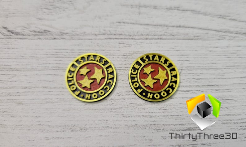 Puede incluir: Dos insignias de metal de color dorado con las palabras "Raccoon City Police Stars" y tres estrellas doradas sobre fondo rojo.
