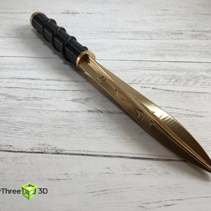 Op de afbeelding: Een decoratieve dolk met een goudkleurig lemmet en een zwarte handgreep. De handgreep heeft een spiraalvormig ontwerp. De dolk ligt op een wit houten oppervlak. Het logo "ThirtyThree 3D" staat in de linker benedenhoek.