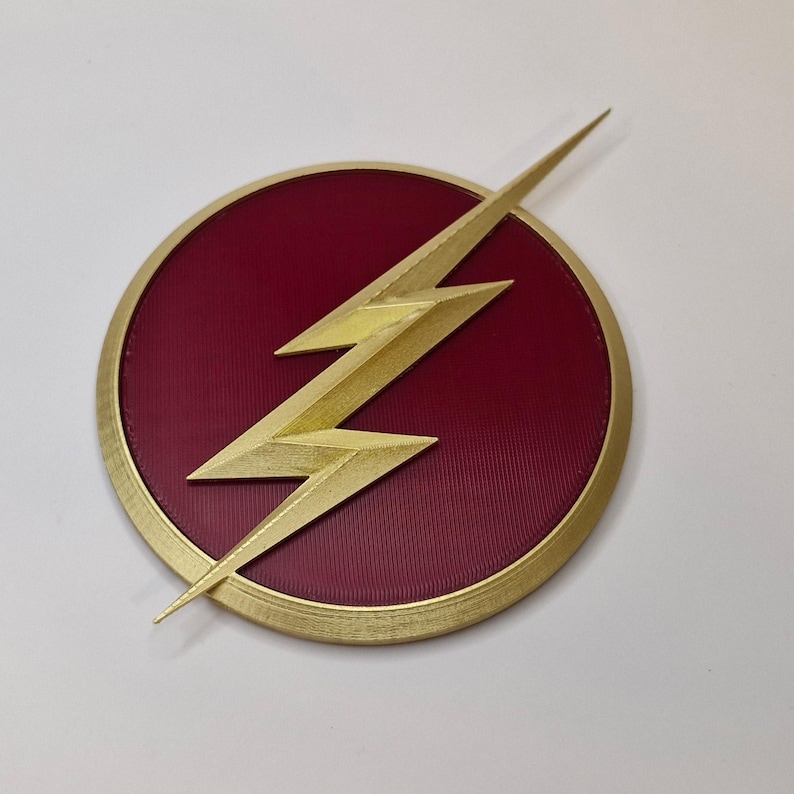 May include: A circular emblem featuring a gold lightning bolt over a maroon background. The lightning bolt is centered within a gold-colored circular border. The maroon background has a textured appearance.