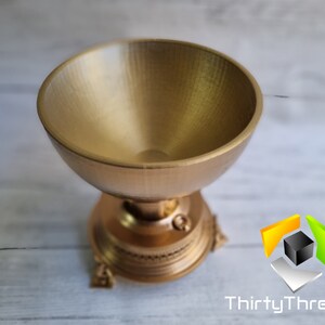 May include: A gold-colored metal trophy with a round, bowl-shaped top and a decorative base. The trophy has a detailed design with intricate carvings.