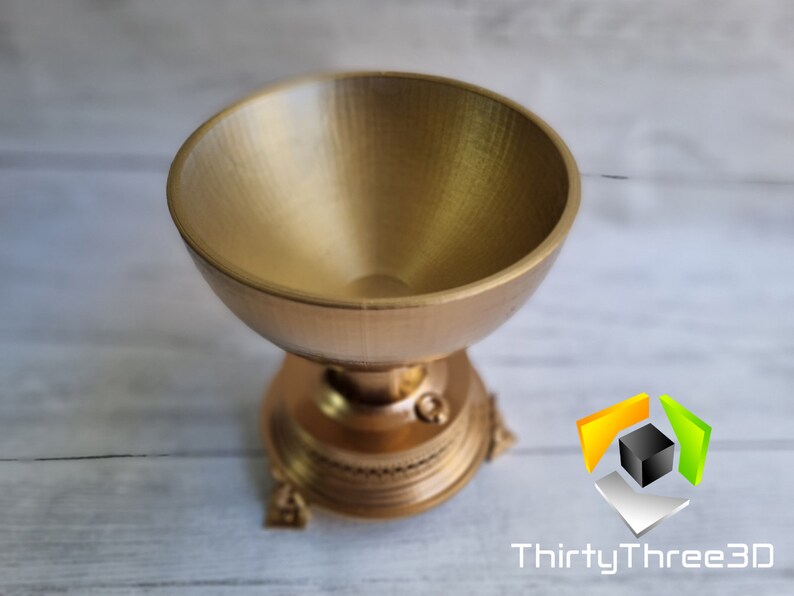 May include: A gold-colored metal trophy with a round, bowl-shaped top and a decorative base. The trophy has a detailed design with intricate carvings.