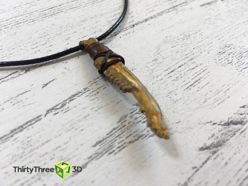 May include: A gold-colored pendant necklace with a bone-shaped charm wrapped in brown leather cord. The necklace is on a black cord.