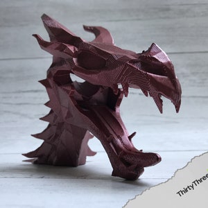 May include: A 3D printed dragon head sculpture in a deep burgundy red color. The sculpture is detailed with sharp teeth and scales.